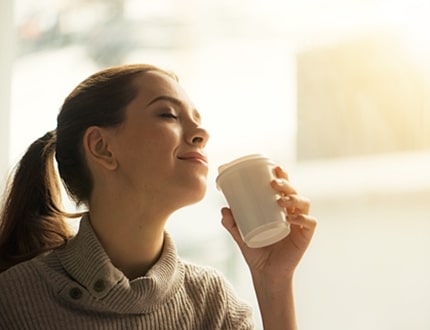 Woman drinking coffee at home with sunrise streaming in through window and creating flare into the lens.